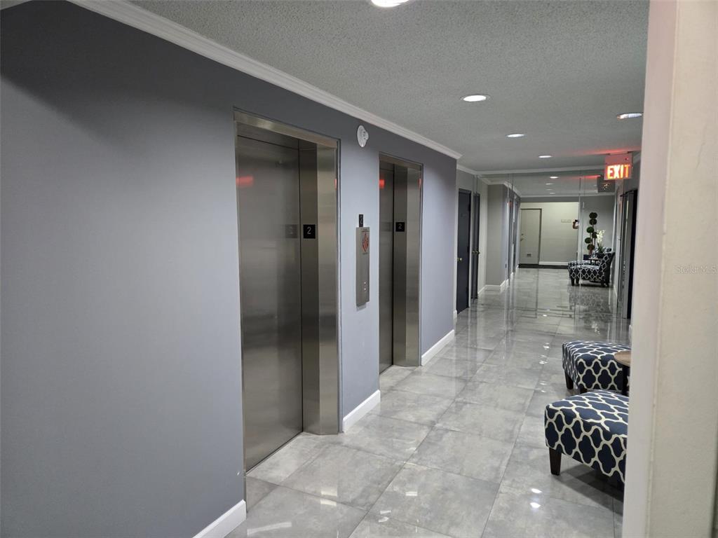 A hallway features two stainless steel elevators, gray walls, and patterned ottomans on tiled flooring — 300 DIPLOMAT PARKWAY UNIT 203, HALLANDALE BEACH, FL