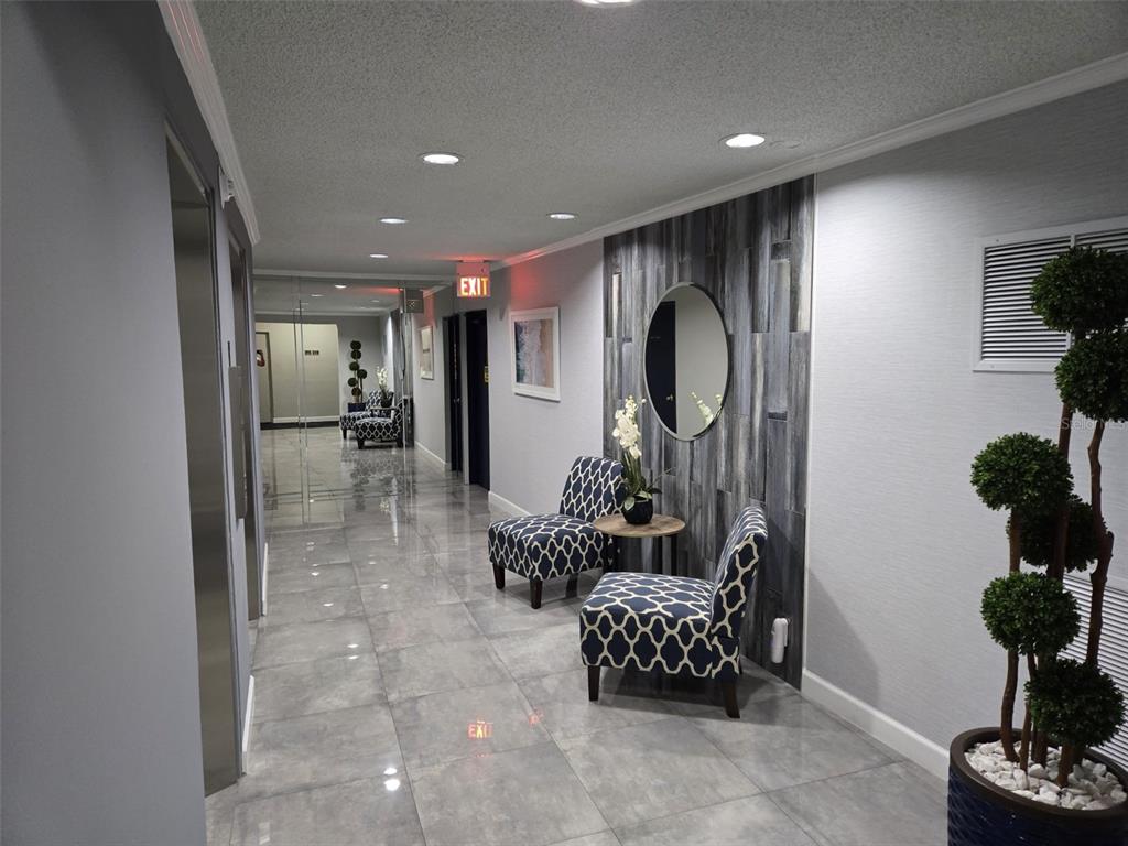 A modern hallway features gray tile flooring, patterned armchairs, a round mirror, and recessed lighting — 300 DIPLOMAT PARKWAY UNIT 203, HALLANDALE BEACH, FL
