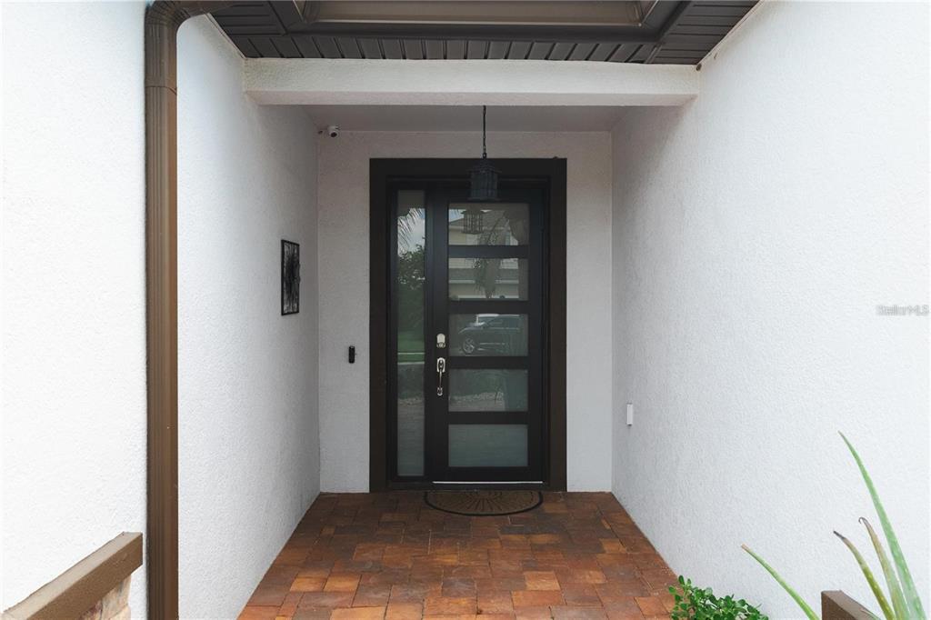 A covered entryway features a dark-framed glass door, a brick-paver floor, and white stucco walls — 5635 FISHER GLEN LOOP, WESLEY CHAPEL, FL