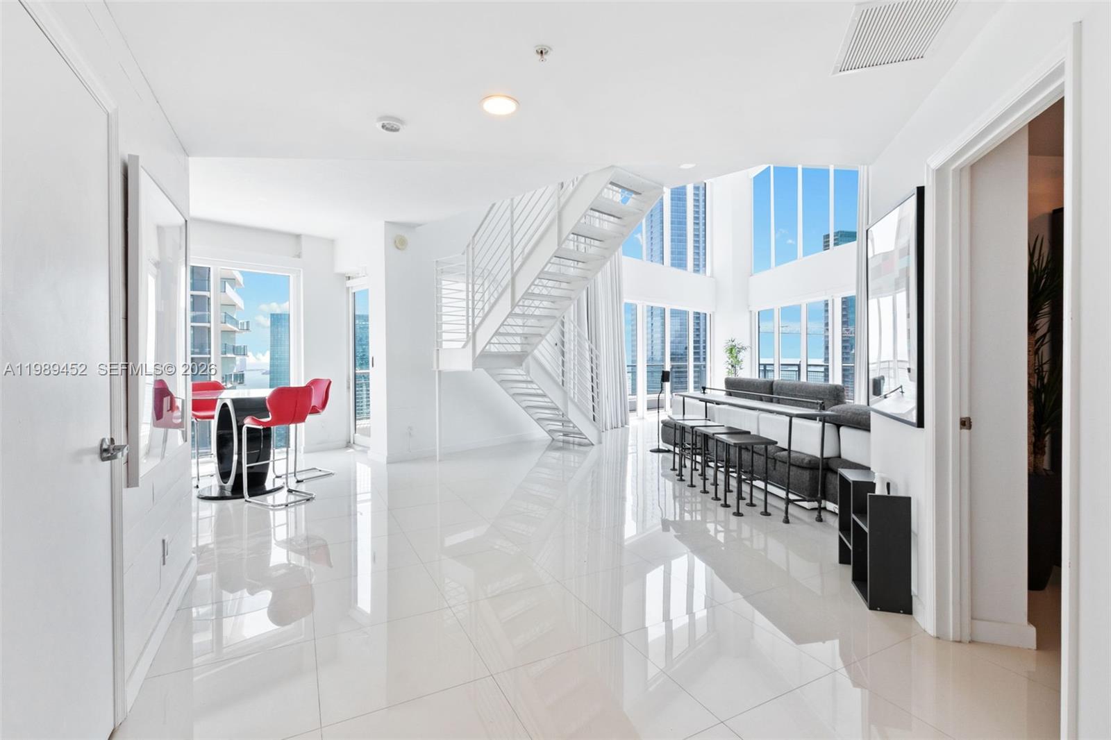A bright, modern living room features white tile floors, a floating staircase, and floor-to-ceiling windows — 60 SW 13th St # 3800, Miami FL 33130, MIAMI, FL