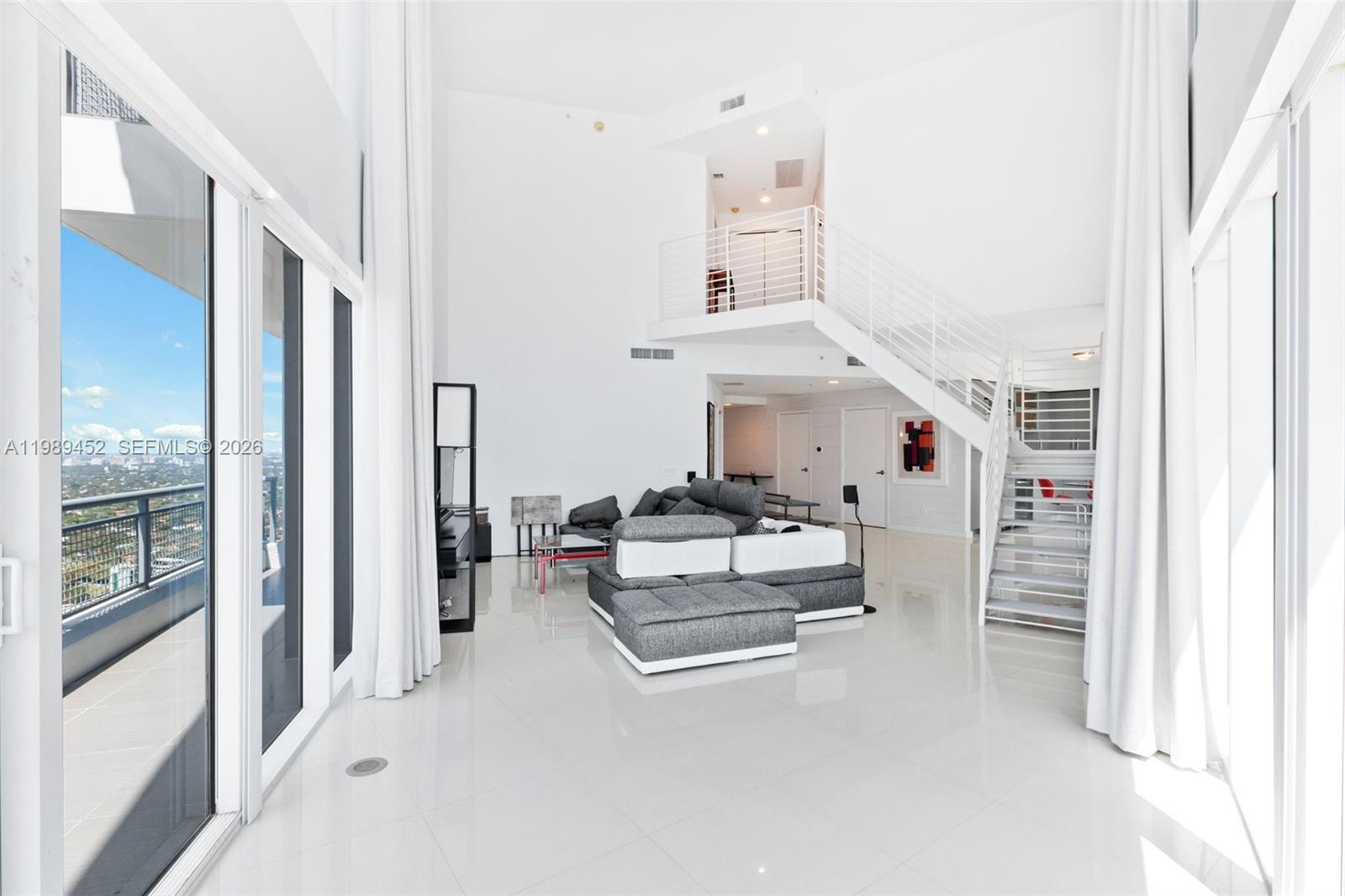 A modern, high-ceilinged living room features white tile floors, a gray sectional, and a staircase — 60 SW 13th St # 3800, Miami FL 33130, MIAMI, FL
