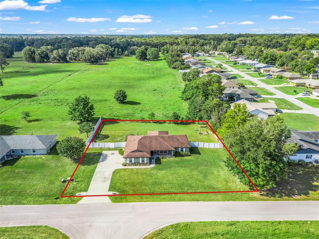 An aerial view shows a single-story house with a fenced backyard bordering a large field — 7474 SE 107 PLACE, BELLEVIEW, FL