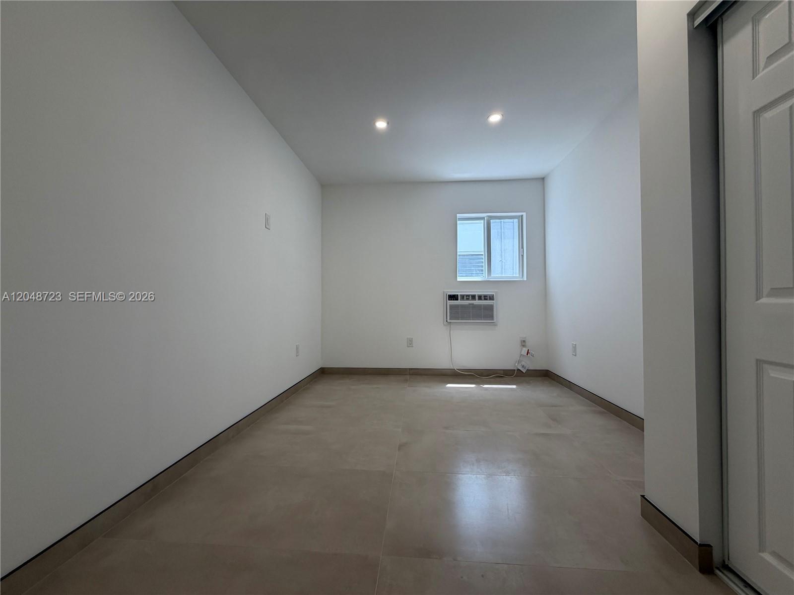 An empty room features white walls, large beige floor tiles, a window, and an air conditioner — 1235 Normandy Dr # 7, Miami Beach FL 33141, MIAMI BEACH, FL