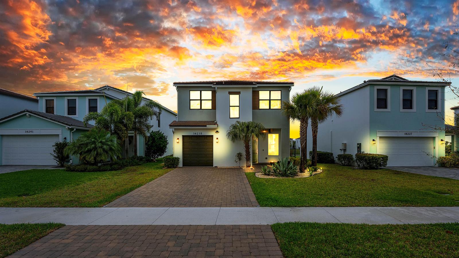 Three suburban houses with paved driveways and lawns stand under a vibrant sunset sky — 16235 Sandy Shore Drive, Westlake FL 33470, WESTLAKE, FL