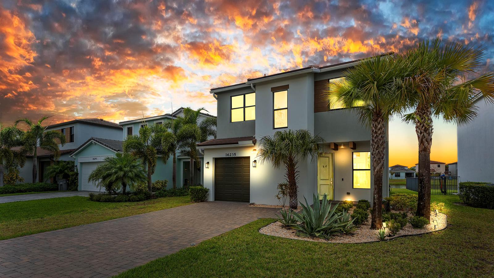 A two-story house with a paved driveway, palm trees, and a sunset sky in the background — 16235 Sandy Shore Drive, Westlake FL 33470, WESTLAKE, FL
