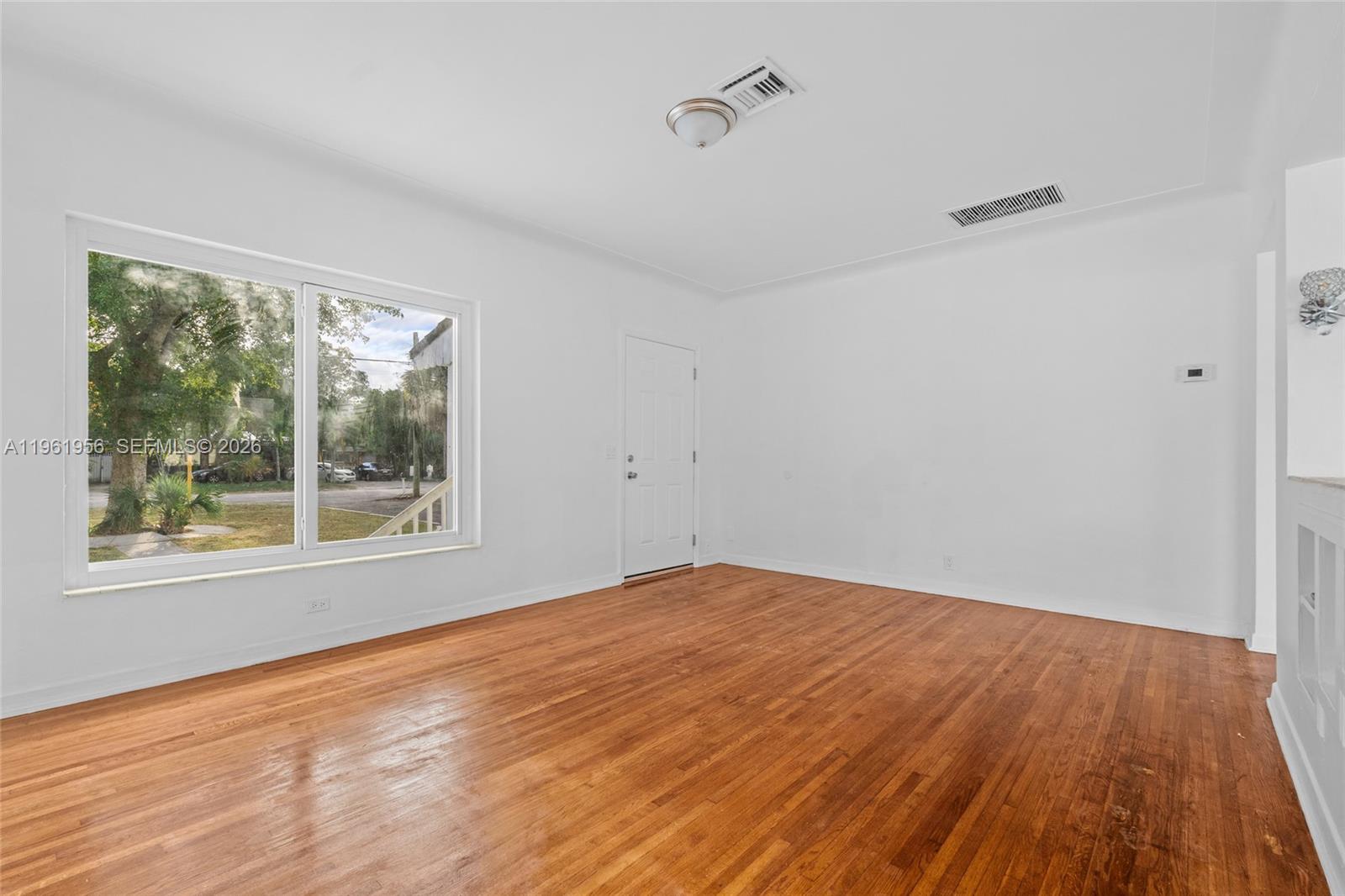 An empty room features white walls, hardwood floors, a large window, and a white door — 201 S Biscayne River Dr, Miami FL 33169, MIAMI, FL
