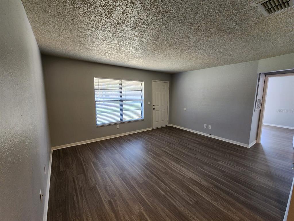 An empty living room features gray walls, wood-look flooring, a window with blinds, and popcorn ceilings — 9530 LYNN LANE UNIT B, SEMINOLE, FL