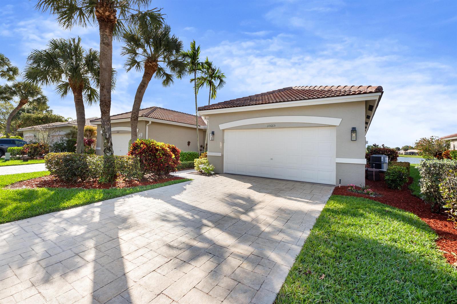 A beige single-story house features a white two-car garage, a paved driveway, and palm trees — 10665 Royal Caribbean Circle, Boynton Beach FL 33437, BOYNTON BEACH, FL