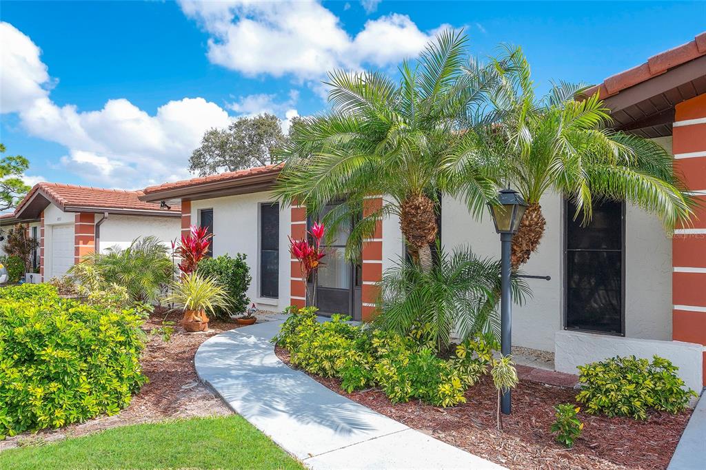 A single-story house exterior features a curved walkway, palm trees, and red-trimmed white walls — 6004 VIVIENDA DRIVE UNIT 8, BRADENTON, FL
