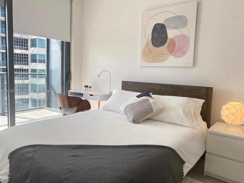 A bedroom features a bed with white bedding, a desk, and a view of buildings — 121 NE 34th St # 1703, Miami FL 33137, MIAMI, FL