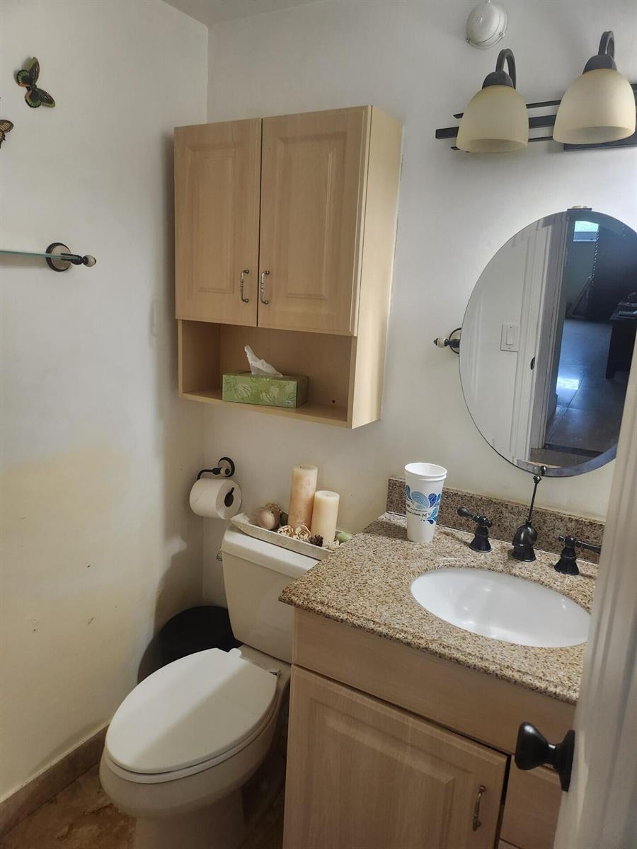 A small bathroom features a granite vanity, light wood cabinets, a round mirror, and toilet — 4200 Hillcrest Drive # 707, Hollywood FL 33021, HOLLYWOOD, FL