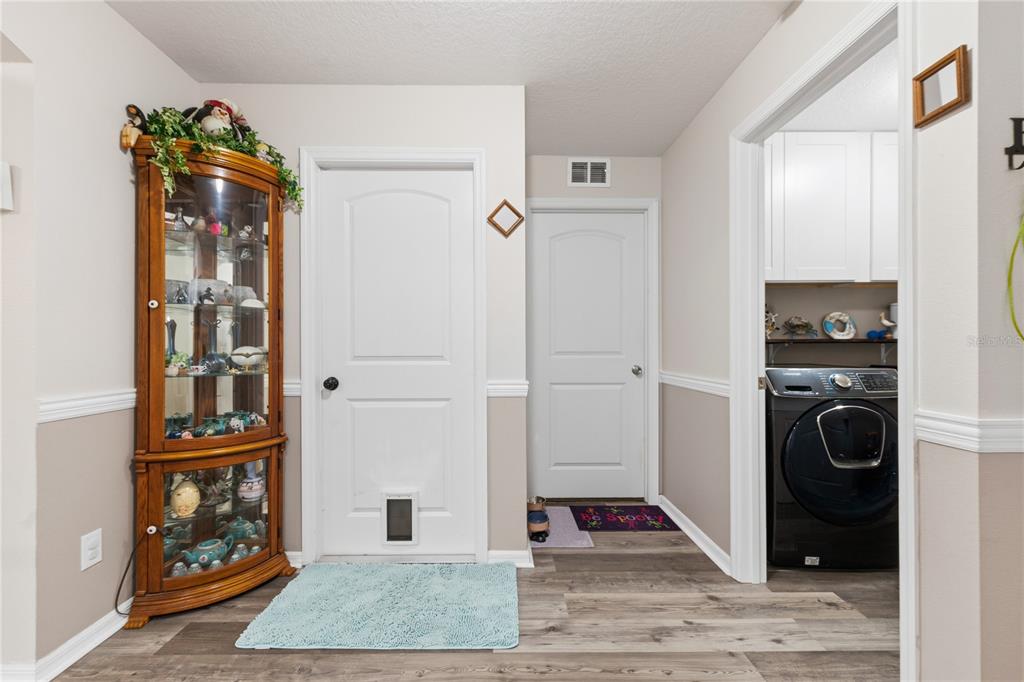 A hallway features wood-look flooring, two white doors, a corner curio cabinet, and a laundry room — 5581 MAGGIORE BOULEVARD, LAKELAND, FL