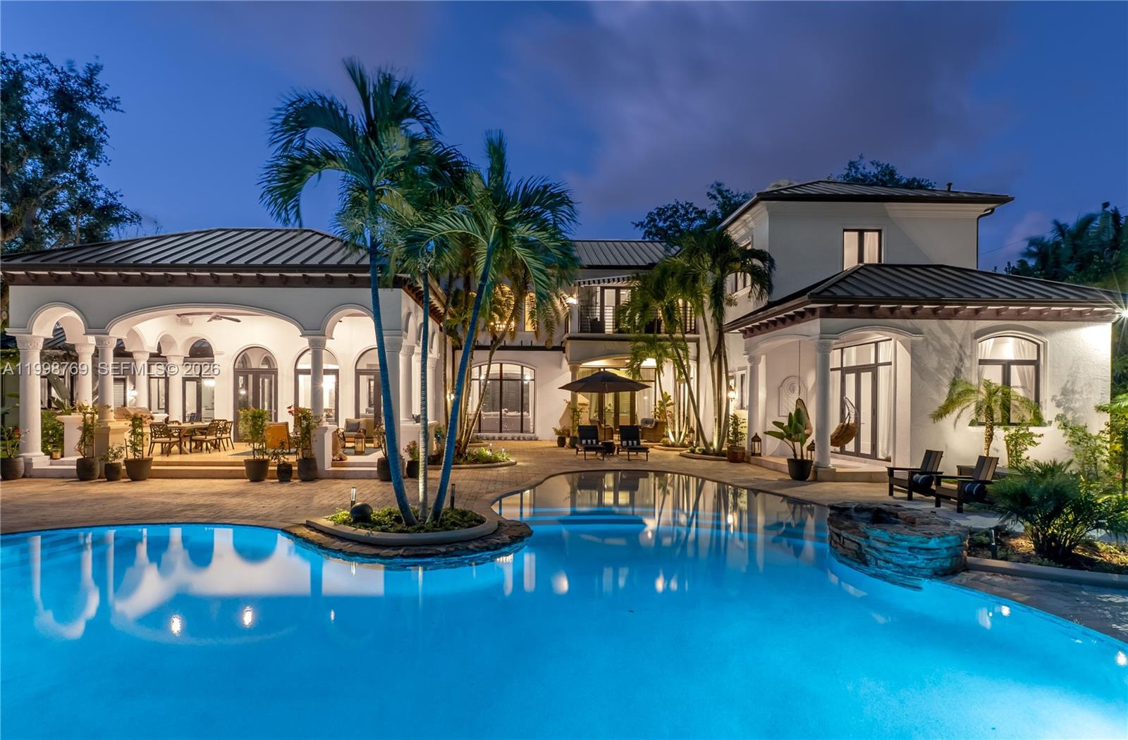 A luxury home exterior at dusk featuring a swimming pool, covered patio, and palm trees — 6350 SW 107th St, Pinecrest FL 33156, PINECREST, FL