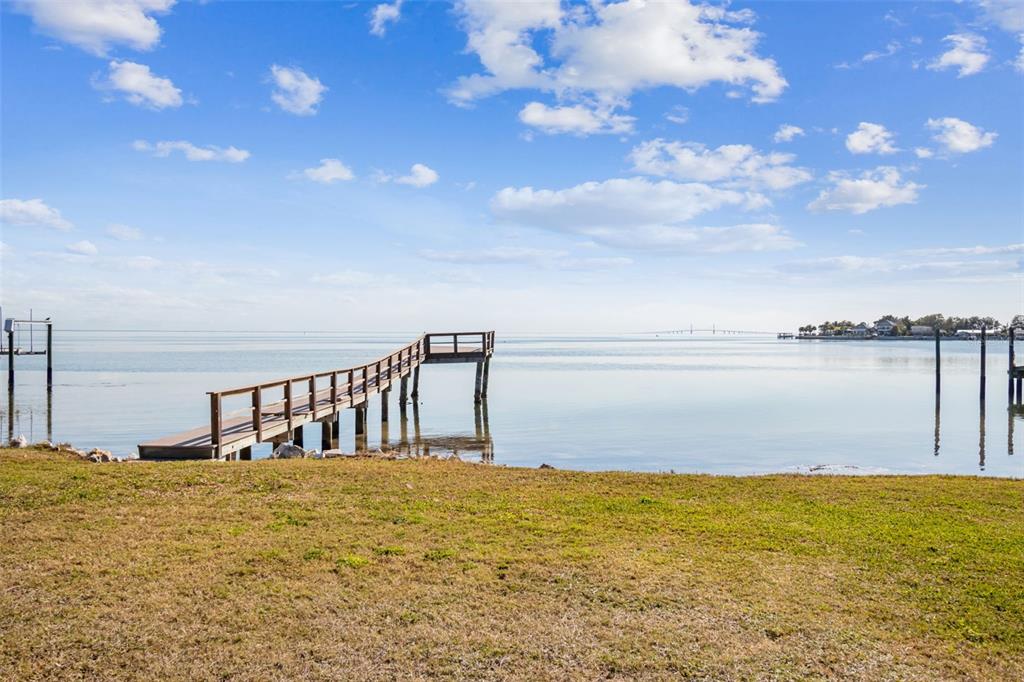 A wooden pier extends into calm water from a grassy shoreline under a blue sky — 6330 BAHAMA SHORES DRIVE S, ST PETERSBURG, FL