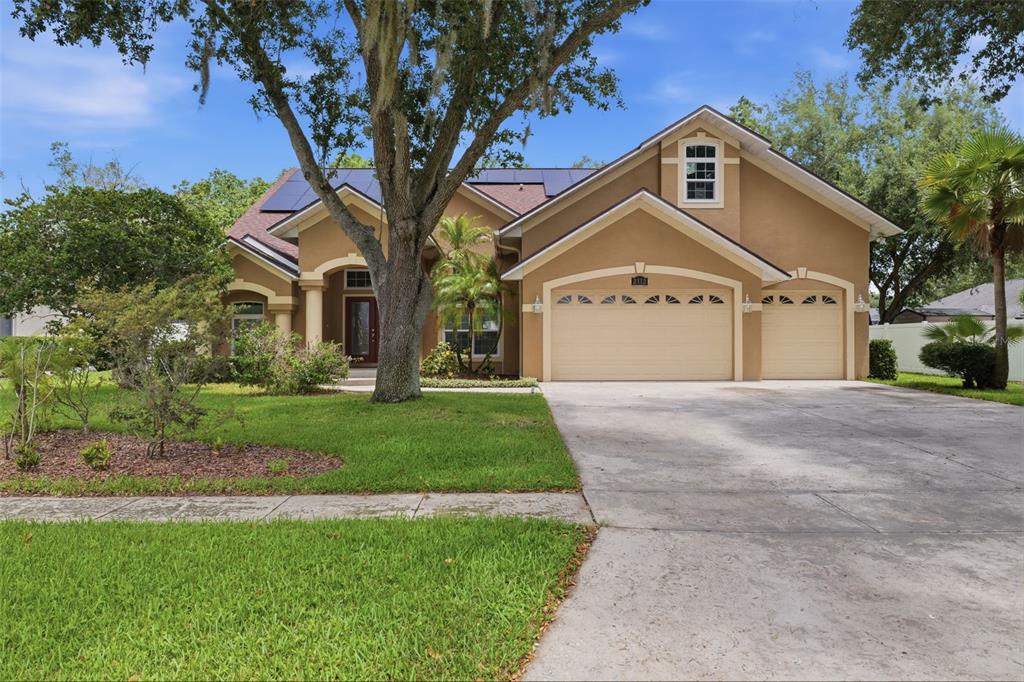 A tan, single-story house with a three-car garage, solar panels, and a large front tree — 2112 HERITAGE CREST DRIVE, VALRICO, FL