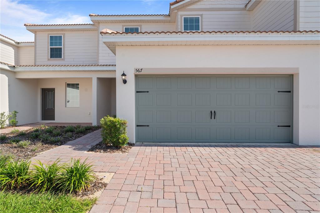 A two-story townhouse exterior features a green garage door, brick driveway, and light-colored siding — 567 BAY LEAF DRIVE, KISSIMMEE, FL