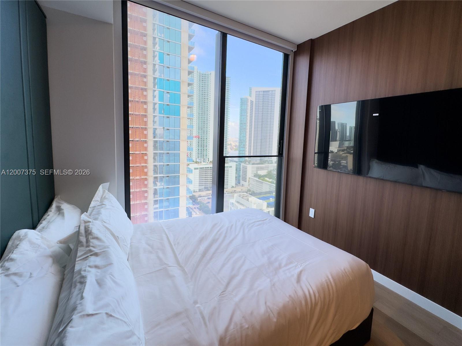 A bedroom features a bed, a wall-mounted television, and floor-to-ceiling windows overlooking city skyscrapers — 20 NE 11th St # 3006B, Miami FL 33132, MIAMI, FL