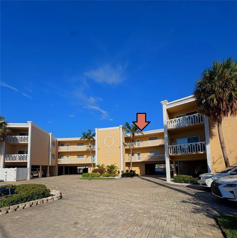 The exterior of a multi-story apartment building features a paved courtyard and palm trees — 11605 3RD STREET E UNIT 306, TREASURE ISLAND, FL