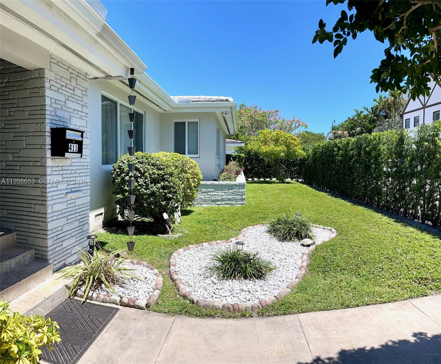 The exterior of a white house features a stone facade, a lawn, and rock landscaping — 411 Alesio Ave, Coral Gables FL 33134, CORAL GABLES, FL