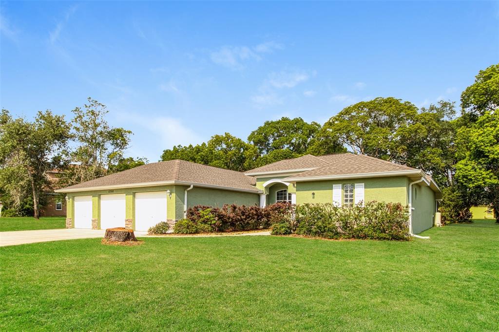 A single-story house with a three-car garage and green stucco exterior sits on a lawn — 13768 RUDI LOOP, SPRING HILL, FL