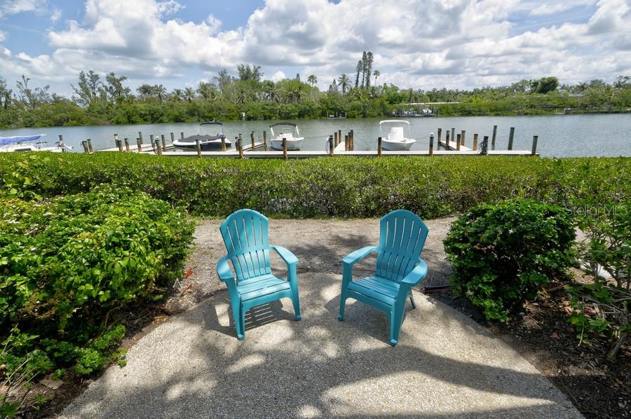 Two blue Adirondack chairs sit on a concrete patio overlooking a canal with boat docks — 6700 GULF OF MEXICO DRIVE UNIT 118, LONGBOAT KEY, FL