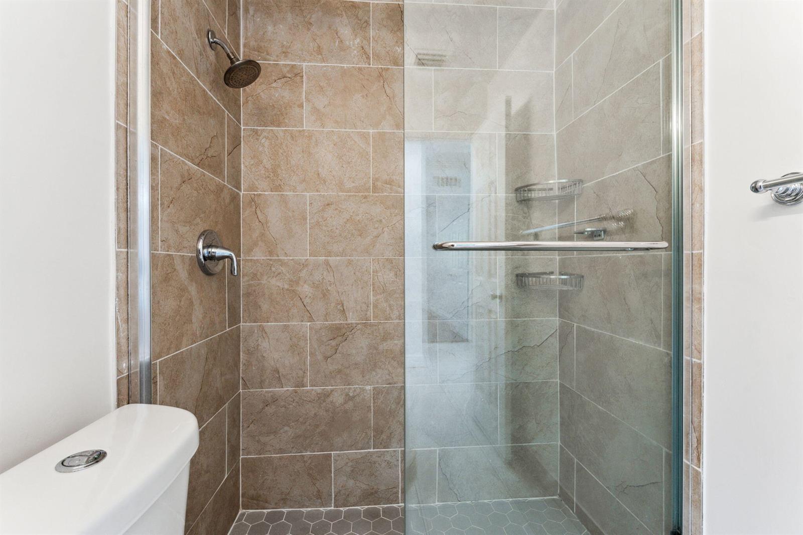 A bathroom shower features tan rectangular wall tiles, a glass door, and hexagonal floor tiles — 441 NW 1st Way, Deerfield Beach FL 33441, DEERFIELD BEACH, FL