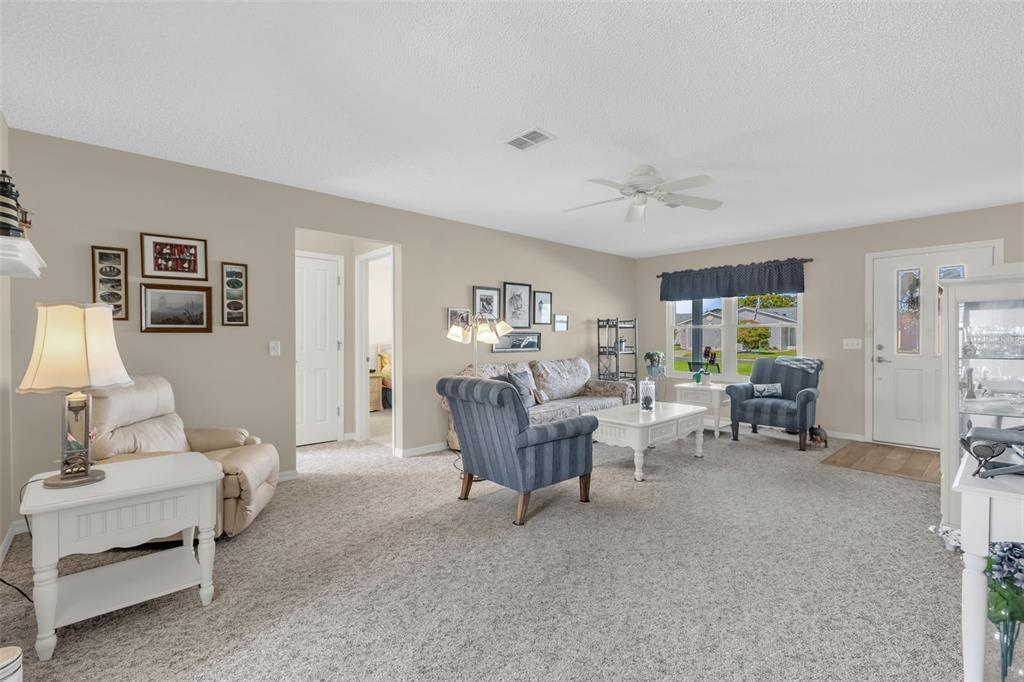 A living room features beige walls, carpeted flooring, a ceiling fan, and various seating arrangements — 1707 GOMEZ PLACE, THE VILLAGES, FL