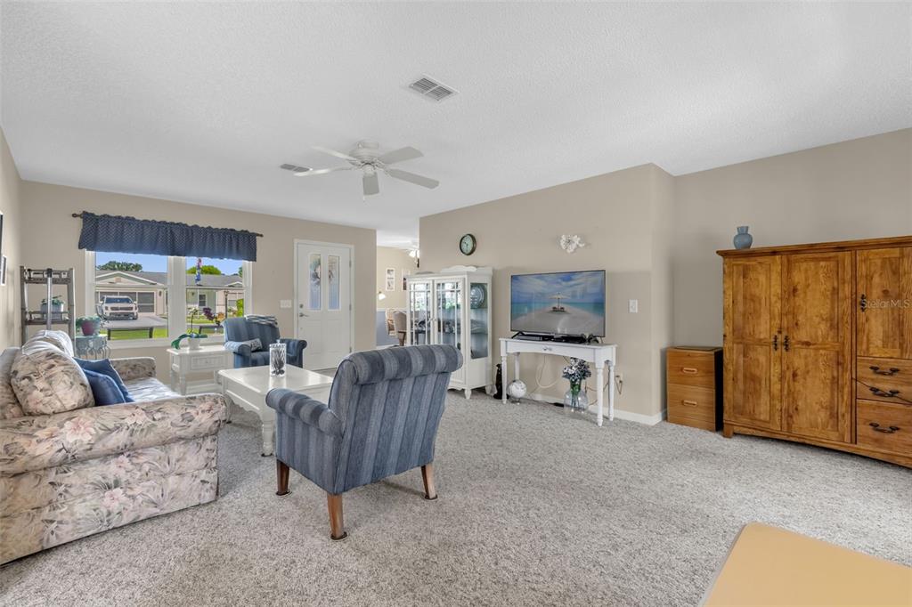 A living room features carpeted flooring, a floral sofa, blue armchairs, and a television stand — 1707 GOMEZ PLACE, THE VILLAGES, FL