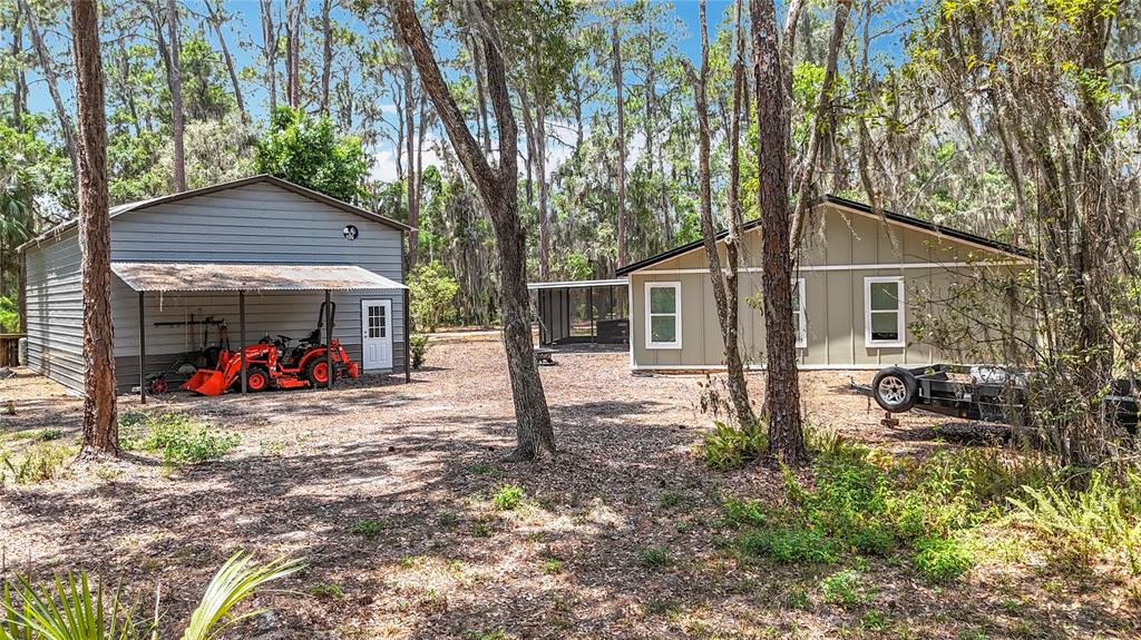 A gray metal workshop and a tan house sit on a wooded, dirt-covered lot — 7374 CIRCLE DRIVE, LADY LAKE, FL