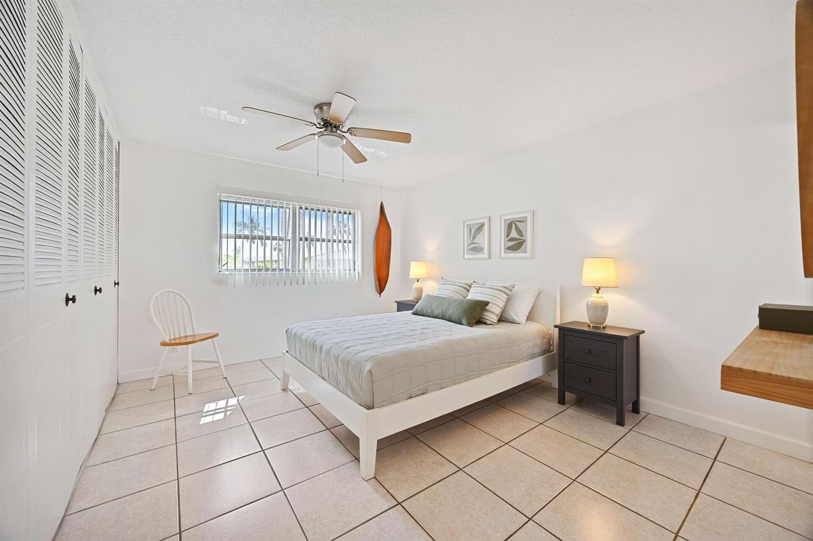 A bedroom features a white bed, tiled floors, a ceiling fan, and white louvered closet doors — 2691 Dudley Dr Drive E # D, West Palm Beach FL 33415, WEST PALM BEACH, FL