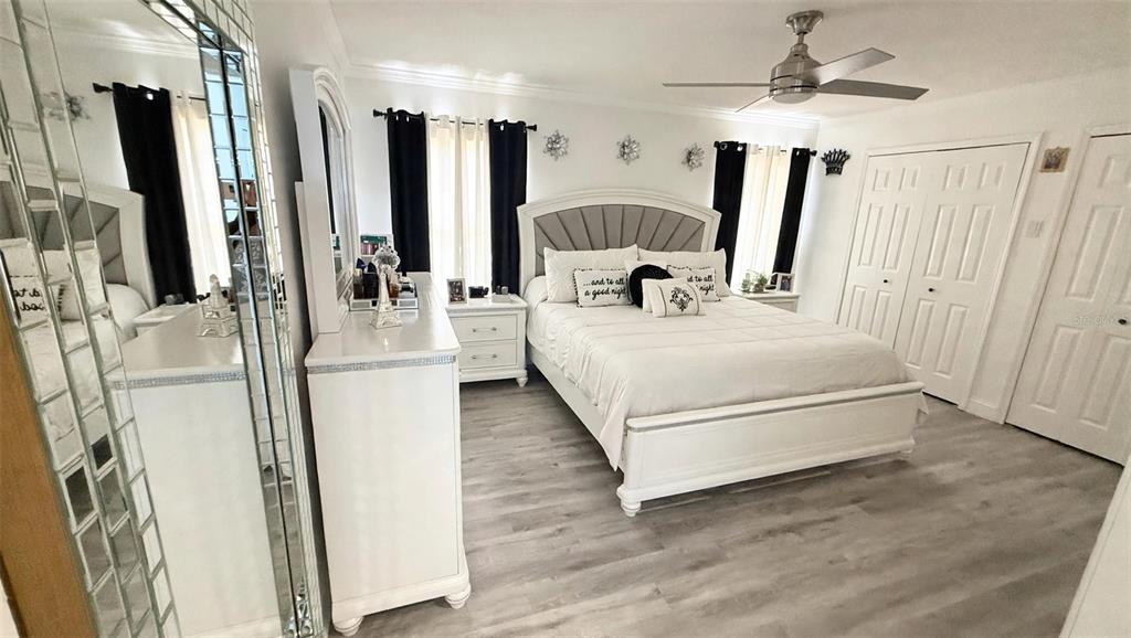 A bedroom features a white bed, matching dresser, mirrored accents, and light wood-look flooring — 7102 HOLLOWELL DRIVE, TAMPA, FL