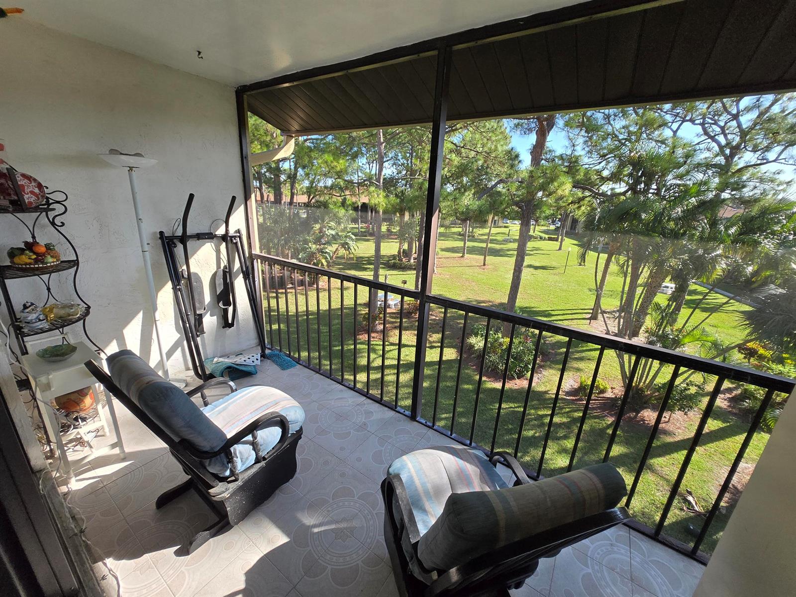 A screened-in balcony features tiled flooring, two rocking chairs, and an exercise machine overlooking trees — 819 Sky Pine Way # G3, FL 33415, FL