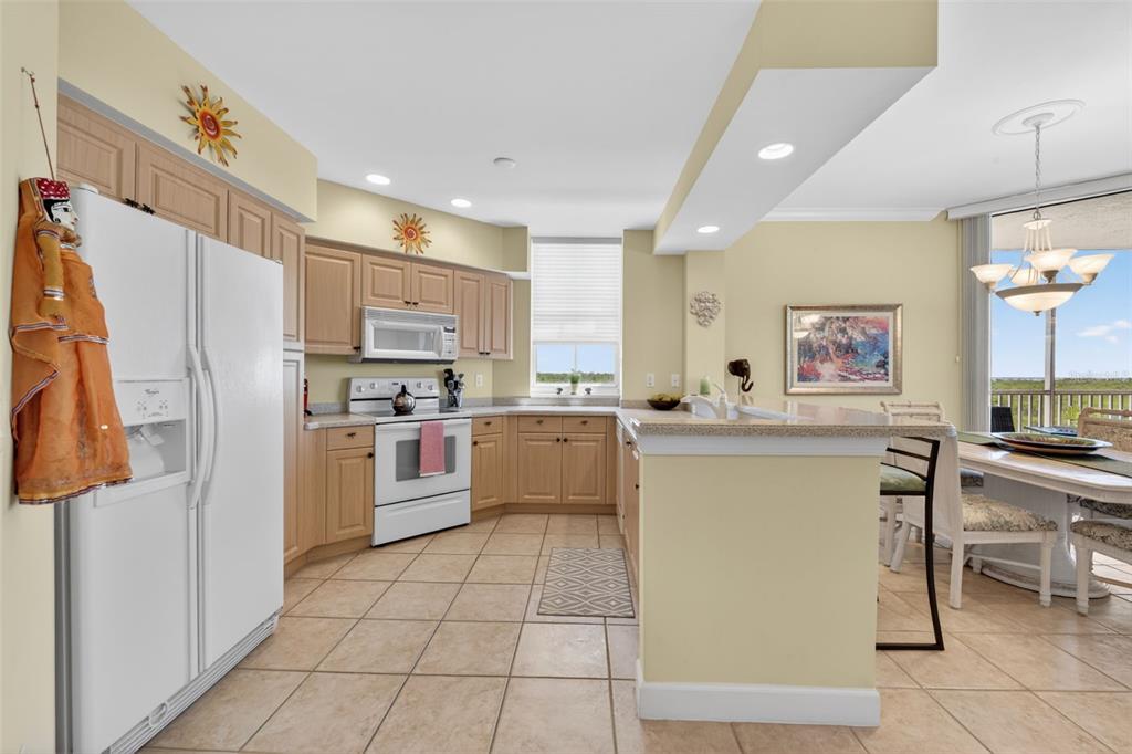 A kitchen with light wood cabinets, white appliances, and tile flooring opens to a dining area — 3191 MATECUMBE KEY ROAD UNIT 202, PUNTA GORDA, FL