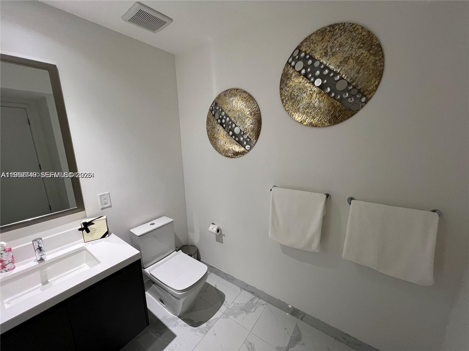 A modern bathroom features a dark vanity, white toilet, marble-look tile flooring, and wall art — 851 NE 1st Ave # 3903, Miami FL 33132, MIAMI, FL