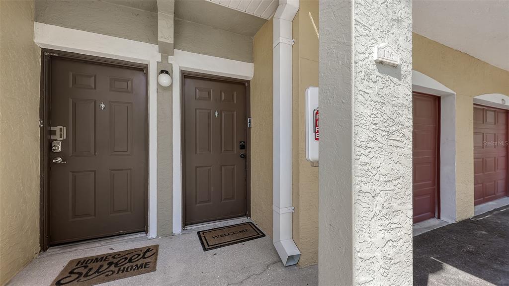 Two brown entry doors with welcome mats are positioned under a covered exterior walkway — 5655 BENTGRASS DRIVE UNIT 9-116, SARASOTA, FL