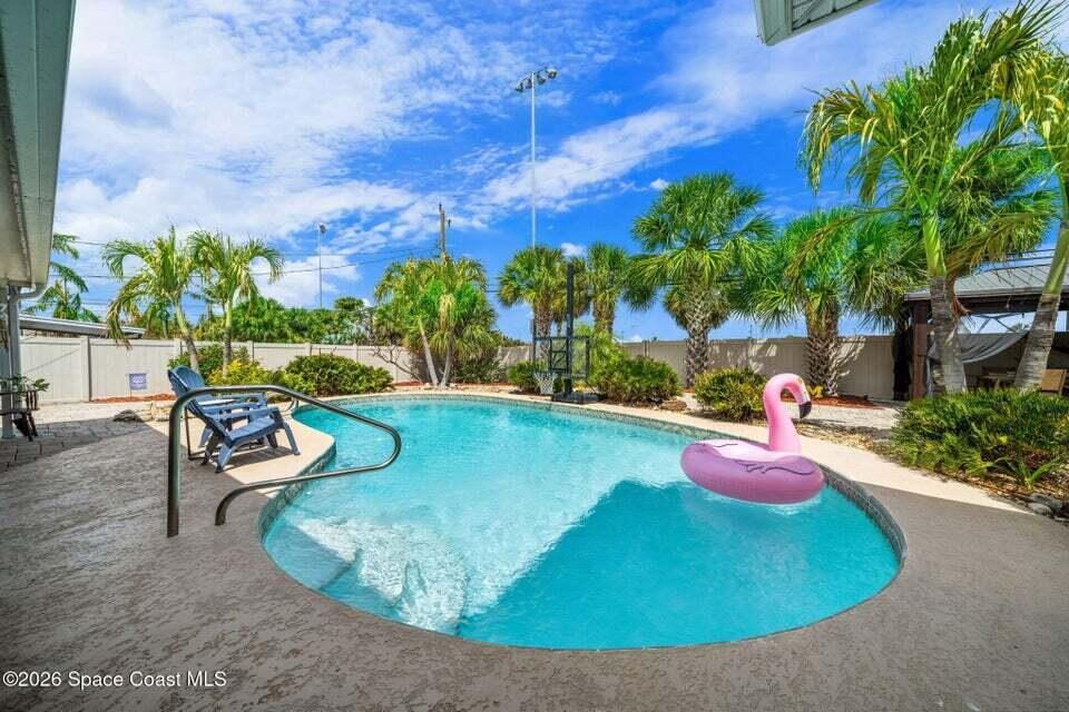 A backyard swimming pool features a pink flamingo float, palm trees, and a privacy fence — 121 Algonquin Terrace, Indian Harbour Beach FL 32937, INDIAN HARBOUR BEACH, FL