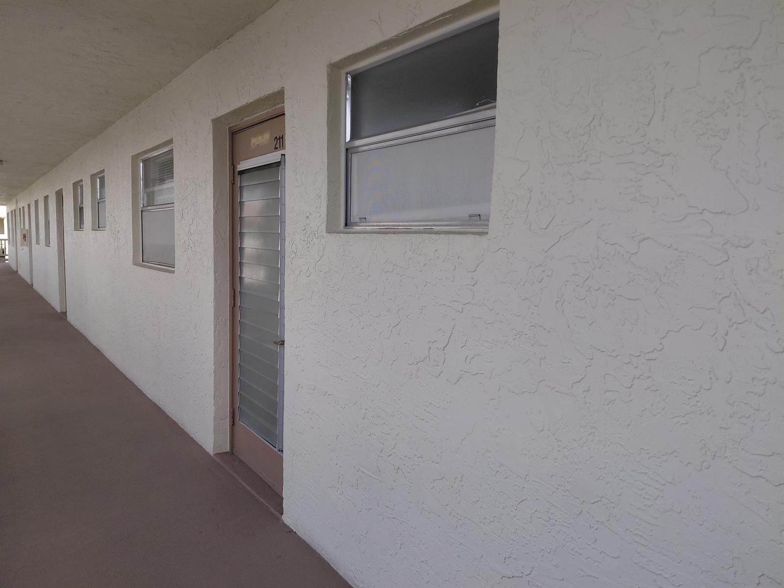 An exterior view of a building hallway featuring white textured walls and louvered doors — 6850 Royal Palm Boulevard # G211, Margate FL 33063, MARGATE, FL