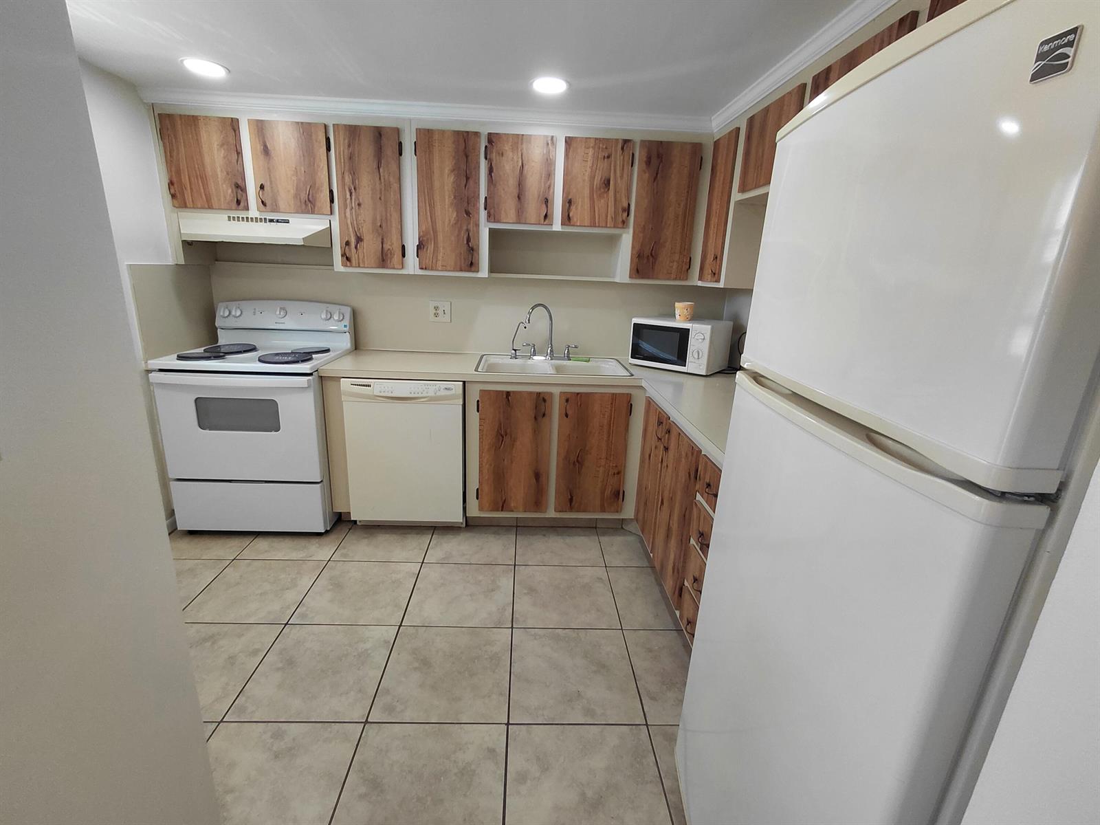 A kitchen features white appliances, wood-grain cabinets, beige countertops, and light-colored tile flooring — 6850 Royal Palm Boulevard # G211, Margate FL 33063, MARGATE, FL