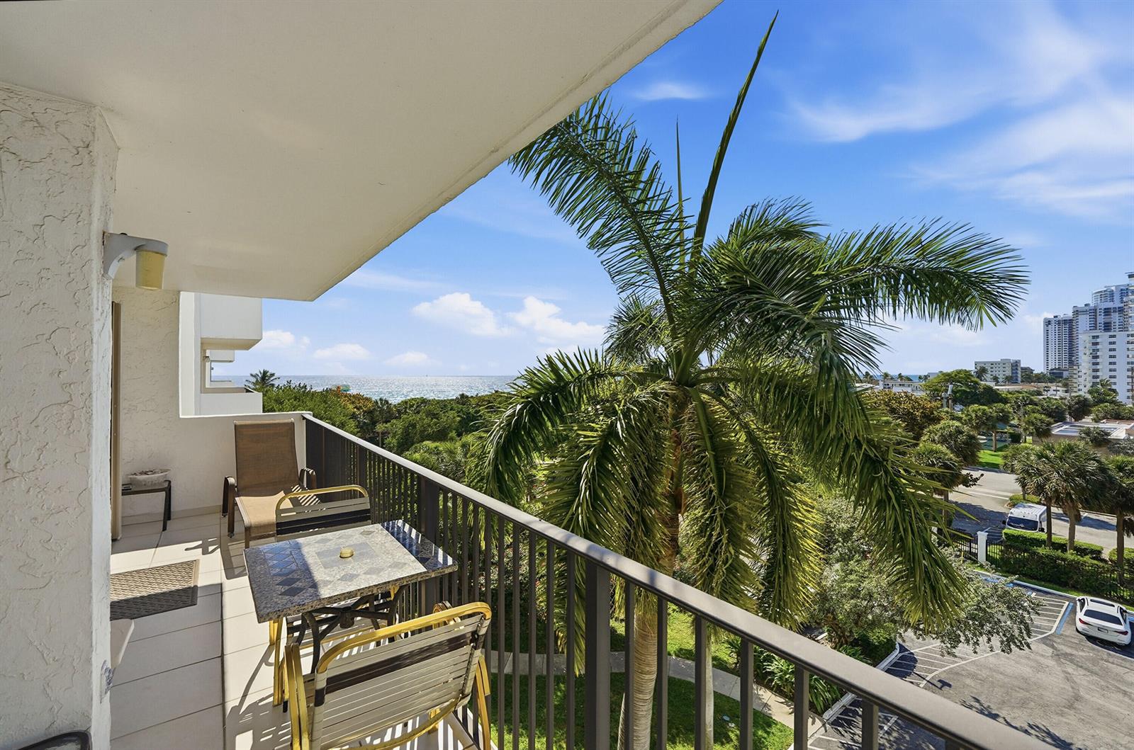 A balcony with patio furniture overlooks a palm tree, lush greenery, and the distant ocean — 1201 S Ocean Drive # 506s, Hollywood FL 33019, HOLLYWOOD, FL