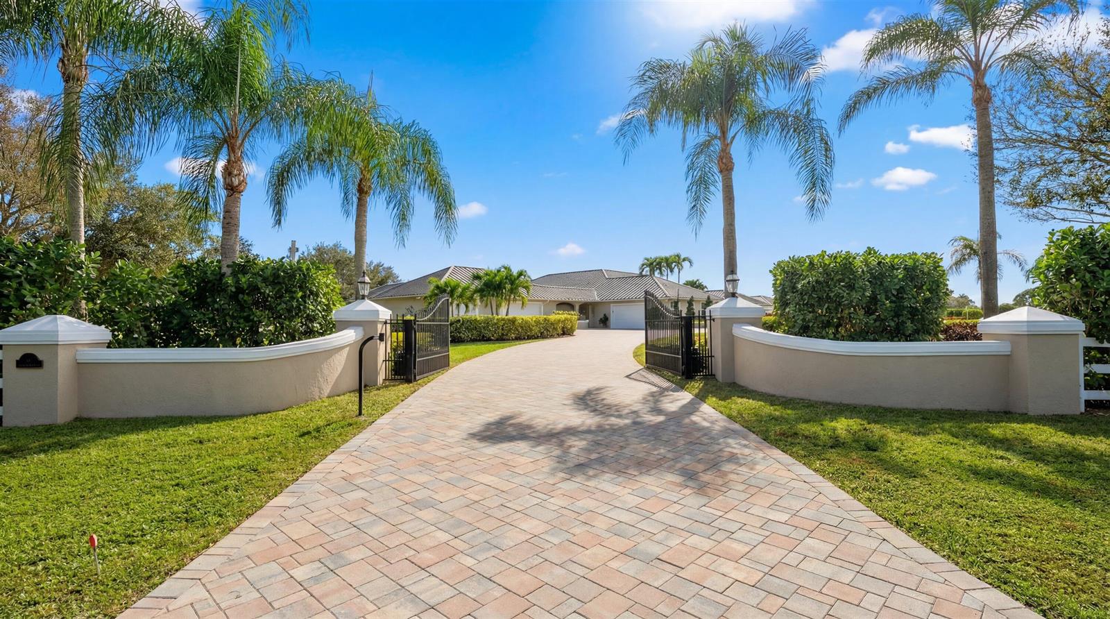 A paved driveway leads through an open gate toward a house with palm trees — 14868 Paddock Drive, Wellington FL 33414, WELLINGTON, FL