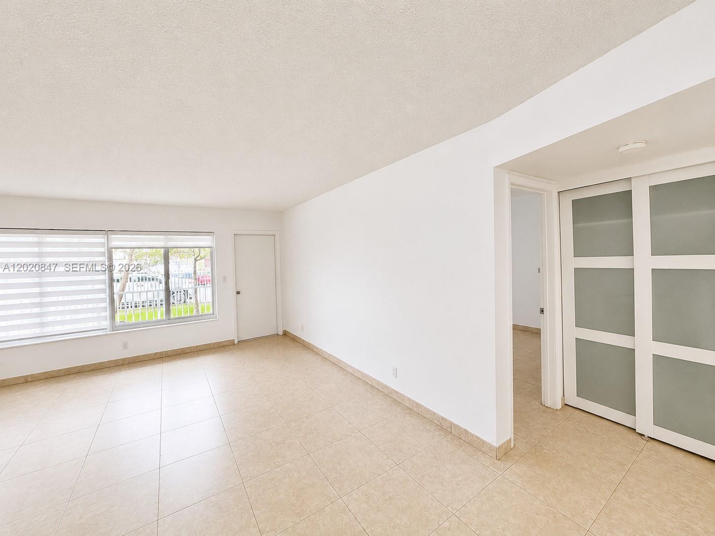 An empty room features white walls, beige tile flooring, a window, and sliding closet doors — 1460 NE 169th St # 108, North Miami Beach FL 33162, NORTH MIAMI BEACH, FL