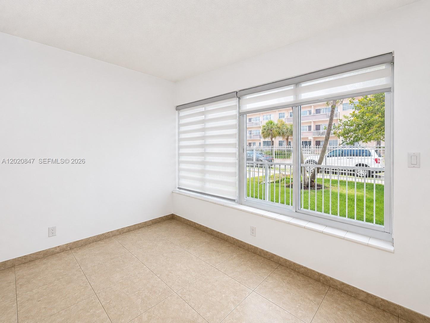 An empty room features beige tile flooring, white walls, and a large window with blinds — 1460 NE 169th St # 108, North Miami Beach FL 33162, NORTH MIAMI BEACH, FL
