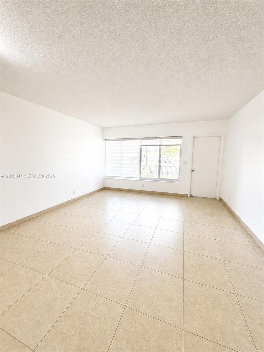 An empty room features light-colored tile flooring, white walls, and a large window — 1460 NE 169th St # 108, North Miami Beach FL 33162, NORTH MIAMI BEACH, FL