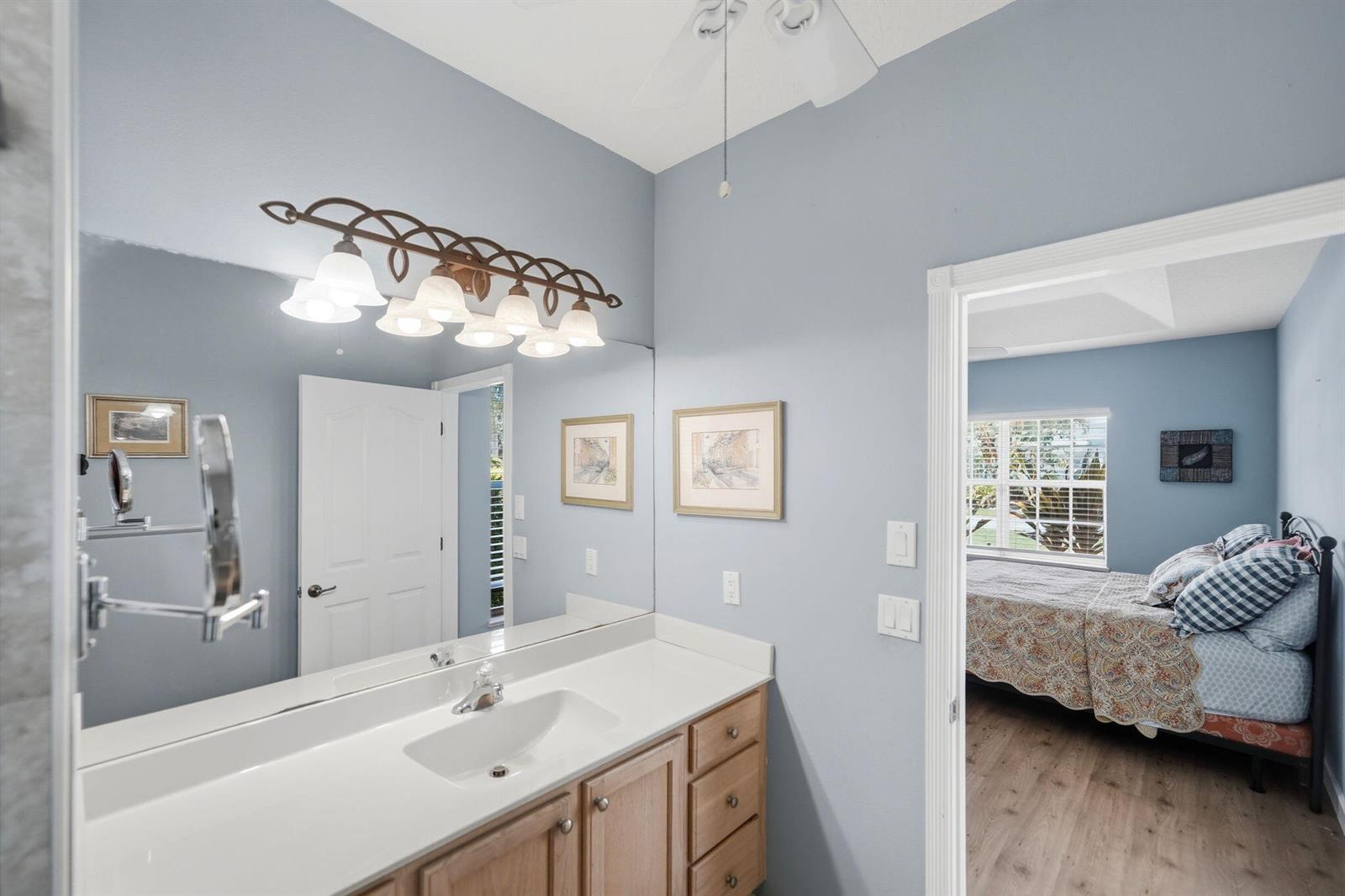 A bathroom with a light wood vanity, white countertop, and mirror, leading into a bedroom — 426 NE Vanda Terrado, Jensen Beach FL 34957, JENSEN BEACH, FL