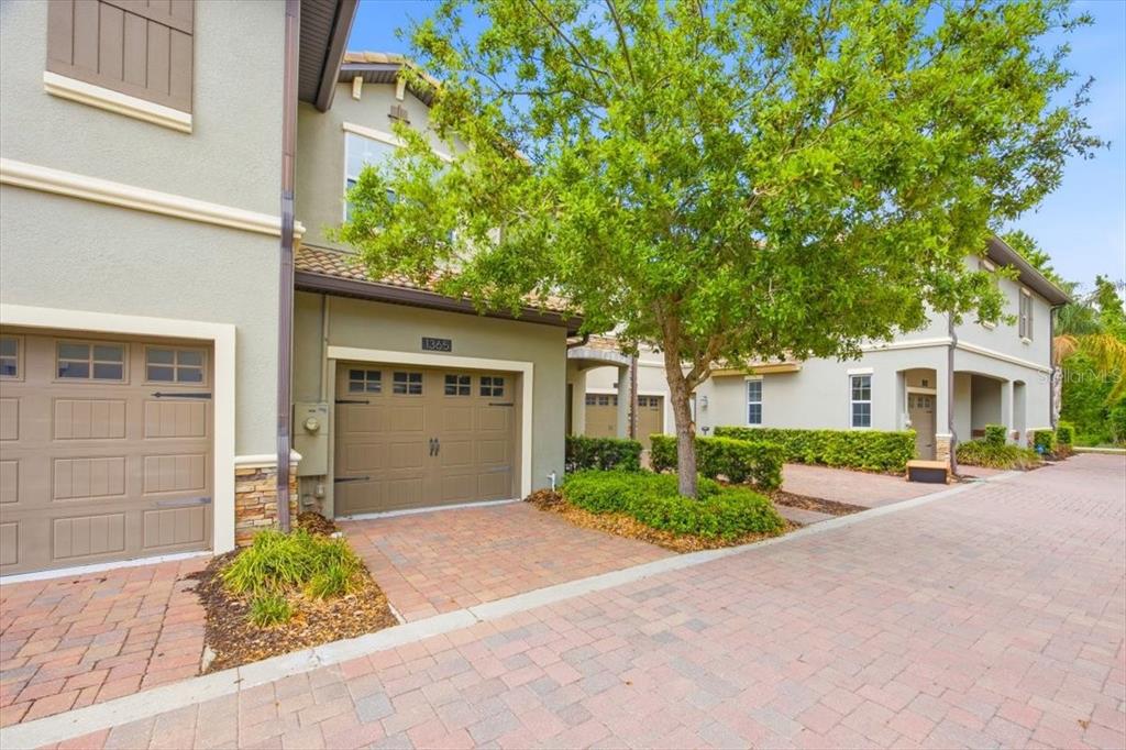 A two-story townhouse exterior with a tan garage, brick driveway, and a large tree — 1365 SHINNECOCK HILLS DRIVE, DAVENPORT, FL
