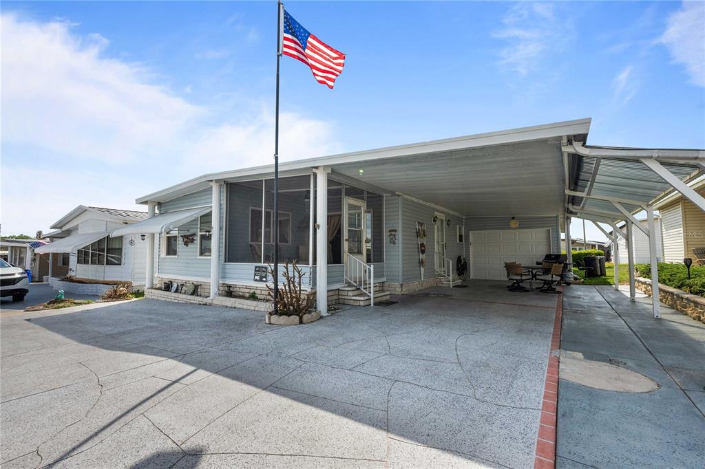 A light blue manufactured home features a screened-in porch, a large carport, and an American flag — 7135 EL DEWITT STREET, ZEPHYRHILLS, FL