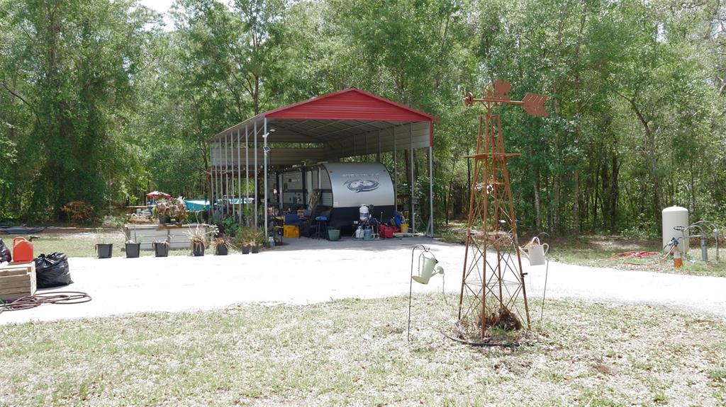 A travel trailer is parked under a metal carport in a wooded yard with a windmill — 17622 CAUFIELD ROAD, SPRING HILL, FL
