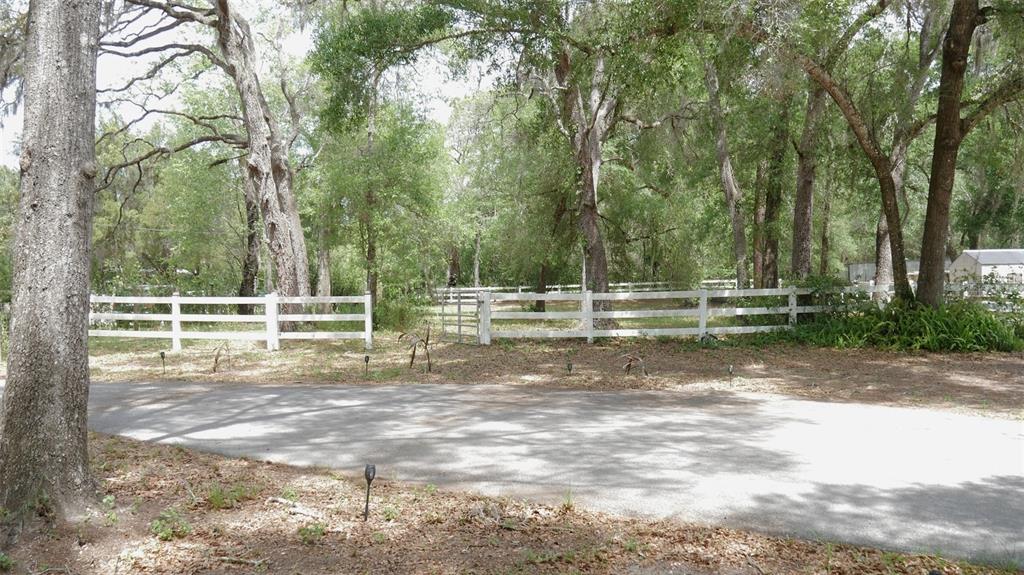 A paved driveway leads to a white wooden fence enclosing a wooded lot with trees — 17622 CAUFIELD ROAD, SPRING HILL, FL