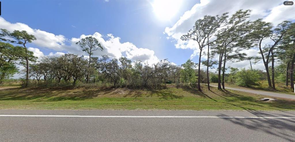 A grassy, wooded vacant lot sits alongside a paved road under a bright, cloudy sky — COMMONWEALTH AVE N, POLK CITY, FL