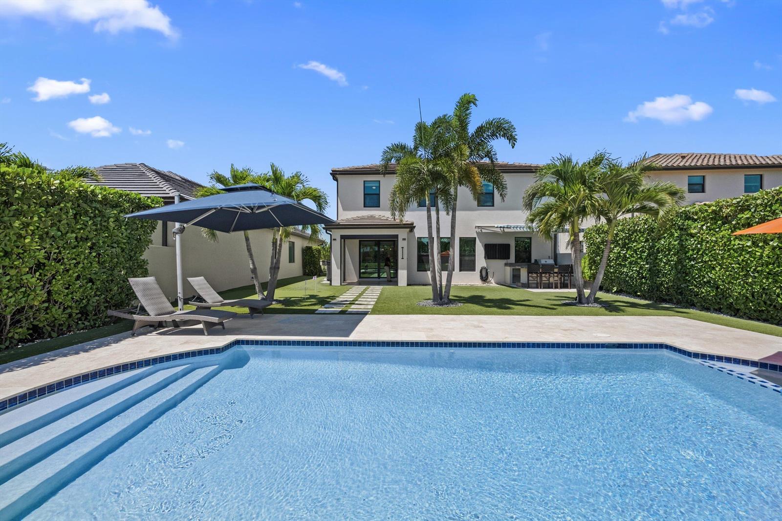 A backyard features a swimming pool, lounge chairs under an umbrella, and palm trees — 8779 Grand Prix Lane, Boynton Beach FL 33472, BOYNTON BEACH, FL
